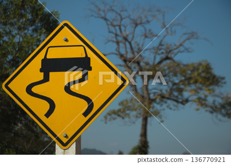 Yellow slippery road warning sign with skidding car symbol on rural roadside under clear blue sky. 136770921