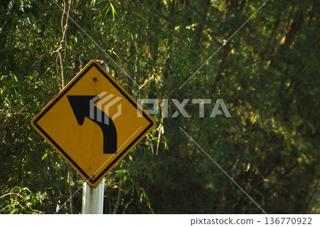 Yellow left curve warning road sign on rural roadside surrounded by green trees in natural daylight. Yellow left curve warning road sign on rural roadside surrounded by green trees in natural daylight. 136770922