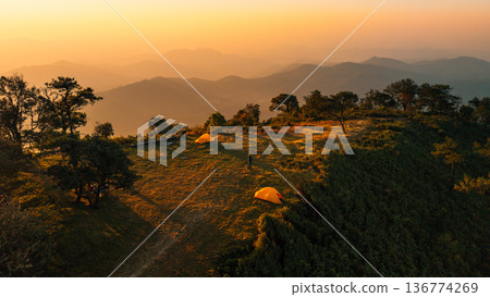 Aerial view of a mountain campsite with yellow tents illuminated by golden morning sunlight. Aerial view of a mountain campsite with yellow tents illuminated by golden morning sunlight. 136774269