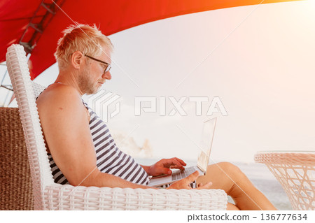 Freelancer laptop beach senior man with glasses working on a laptop while sitting under a red umbrella at a tropical resort for remote work 136777634