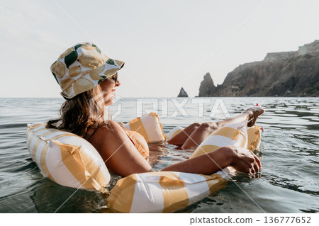 Woman Inflatable Raft Sea Relaxation - A woman wearing a hat and sunglasses relaxes on an inflatable raft in the sea, with rocky cliffs in the background. 136777652
