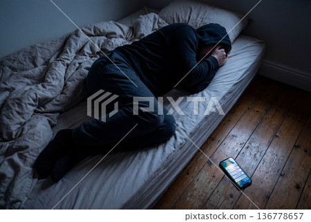 Depressed young caucasian male lying in bed with smartphone nearby in dim room 136778657