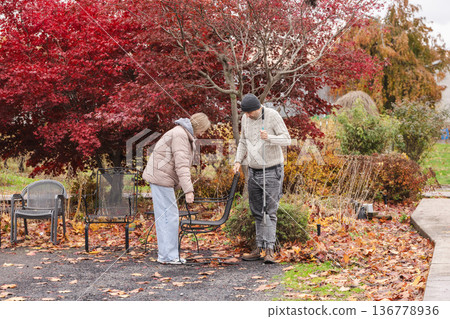 Father and teenage daughter preparing outdoor cafe terrace for winter season while cleaning fallen autumn leaves together 136778936