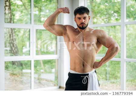 Shows the muscles. Shirtless strong man is standing against panoramic windows Shows the muscles. Shirtless strong man is standing against panoramic windows 136779394