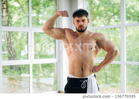Shows the muscles. Shirtless strong man is standing against panoramic windows Shows the muscles. Shirtless strong man is standing against panoramic windows 136779395