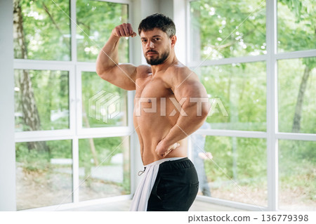 Shows the muscles. Shirtless strong man is standing against panoramic windows Shows the muscles. Shirtless strong man is standing against panoramic windows 136779398