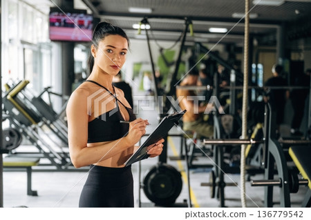 With notepad. Female trainer is standing in the gym With notepad. Female trainer is standing in the gym 136779542