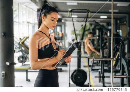 With notepad. Female trainer is standing in the gym With notepad. Female trainer is standing in the gym 136779543