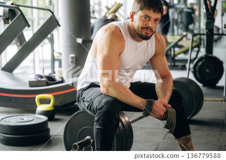 Taking a break, sitting on the barbell. Strong man is working out in the gym Taking a break, sitting on the barbell. Strong man is working out in the gym 136779588