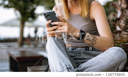 Woman sits outside while using smartphone in a casual setting during daytime Woman sits outside while using smartphone in a casual setting during daytime 136779845