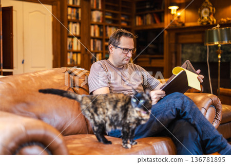 Man reading notebook while sitting on sofa at home with cat in cozy living room Man reading notebook while sitting on sofa at home with cat in cozy living room 136781895