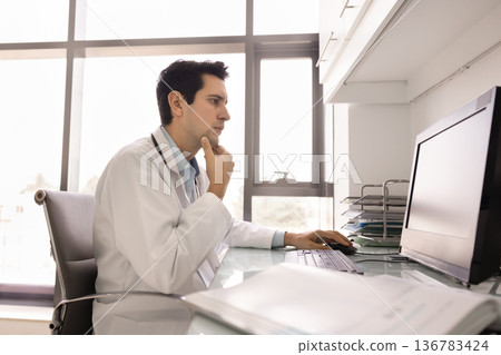 Young 30s male doctor sits at desk working on computer Young 30s male doctor sits at desk working on computer 136783424