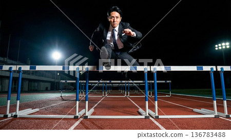 商業圖：一名身穿西裝的男子在夜間的體育場內躍過障礙物。 136783488