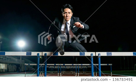 商業圖：一名身穿西裝的男子在夜間的體育場內躍過障礙物。 136783491