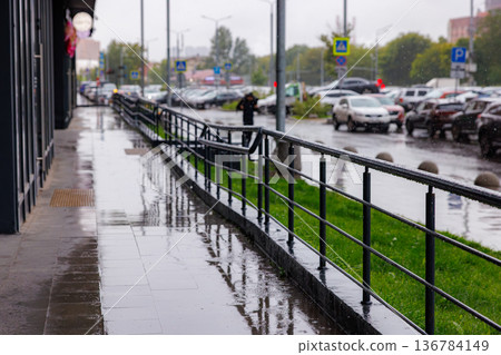 Rain-soaked sidewalk with reflection and parked cars in urban setting during cloudy day 136784149