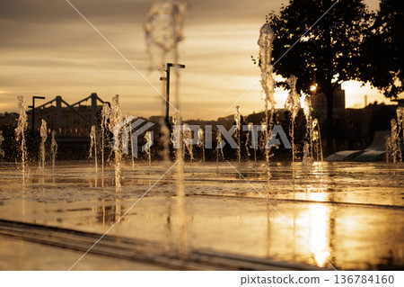 Evening glow of water fountains dancing in the warm sunset light by the riverside promenade Evening glow of water fountains dancing in the warm sunset light by the riverside promenade 136784160