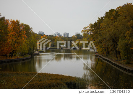 Autumn hues reflect in calm water of park lake surrounded by city skyline in afternoon light Autumn hues reflect in calm water of park lake surrounded by city skyline in afternoon light 136784350