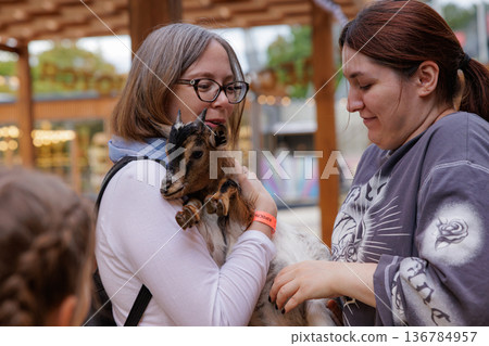 Smiles and laughter as women bond with a playful baby goat at a local petting zoo Smiles and laughter as women bond with a playful baby goat at a local petting zoo 136784957