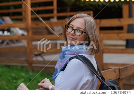 Smiling woman enjoying outdoor event at cozy venue during warm evening, surrounded by wooden decor Smiling woman enjoying outdoor event at cozy venue during warm evening, surrounded by wooden decor 136784974