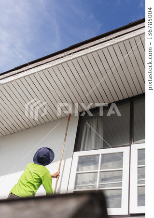 Worker painting exterior house roof with a roller during home renovation. 136785004