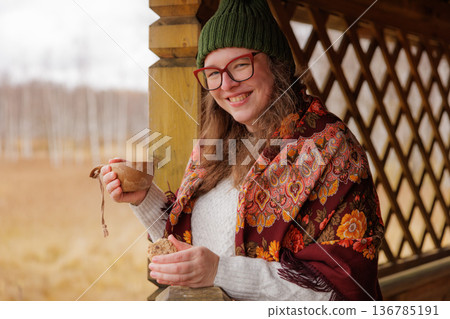 Smiling woman wearing a colorful scarf enjoys tea at a wooden gazebo in a serene autumn setting 136785191