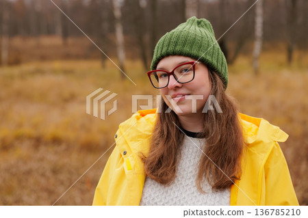 Bright yellow raincoat worn by a cheerful young woman in a serene autumn landscape 136785210