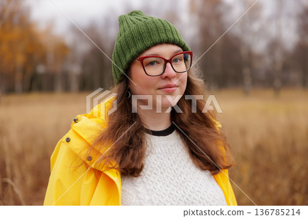 Young woman enjoys a chilly day in a bright yellow jacket and warm green beanie while standing in a field 136785214