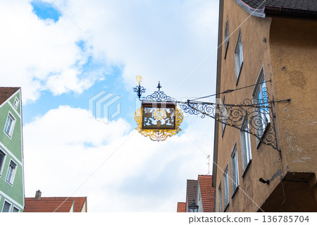 A decorative metal bracket hangs above colorful houses in Memmingen, Germany. The scene features vibrant architecture under a blue sky with clouds. A decorative metal bracket hangs above colorful houses in Memmingen, Germany. The scene features vibrant architecture under a blue sky with clouds. 136785704