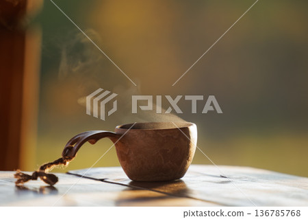 Warm cup of steaming coffee rests on a rustic wooden table in the warm morning sunlight Warm cup of steaming coffee rests on a rustic wooden table in the warm morning sunlight 136785768