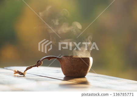 Warm steam floats from a rustic cup in a serene outdoor setting at dawn 136785771