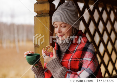 Enjoying a warm drink and cookie while wrapped in a cozy blanket in a serene outdoor setting 136785820