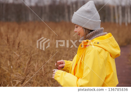 Bright yellow raincoat glows in the autumn field as a woman collects wild grasses amidst nature's palette Bright yellow raincoat glows in the autumn field as a woman collects wild grasses amidst nature's palette 136785836