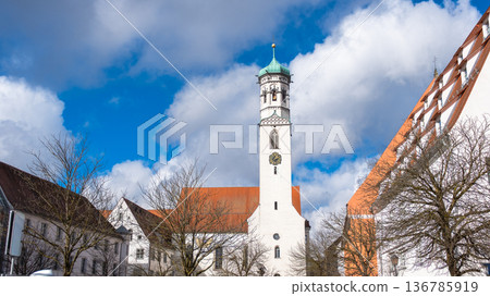 The Hospital Church of St. Peter and Paul in Memmingen, Germany, features a tall clock tower, surrounded by trees and traditional buildings under a blue sky with clouds. The Hospital Church of St. Peter and Paul in Memmingen, Germany, features a tall clock tower, surrounded by trees and traditional buildings under a blue sky with clouds. 136785919