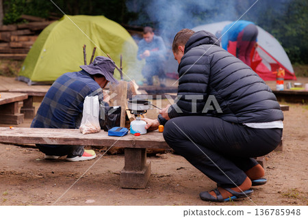 Campers gather around the fire, preparing a warm meal in a serene outdoor setting Campers gather around the fire, preparing a warm meal in a serene outdoor setting 136785948