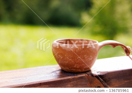 Handcrafted clay cup rests on wooden railing, catching morning sunlight amidst lush greenery Handcrafted clay cup rests on wooden railing, catching morning sunlight amidst lush greenery 136785961