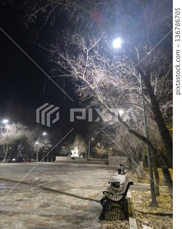 A serene night scene in a park with bare trees and glowing streetlights. Benches offer rest, while a distant statue adds charm, ideal for tourism or urban lifestyle themes. 136786705
