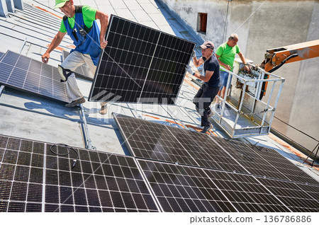 Workers building solar panel system on metal rooftop of house with assistance of crane lift. Two men installers carrying photovoltaic solar module outdoors. Renewable energy generation concept. Workers building solar panel system on metal rooftop of house with assistance of crane lift. Two men installers carrying photovoltaic solar module outdoors. Renewable energy generation concept. 136786886