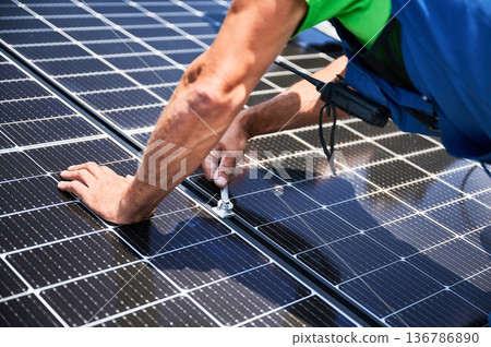 Worker building photovoltaic solar panel system on rooftop of house. Close up of man engineer installing solar module with help of wrench outdoors. Alternative and renewable energy generation concept. 136786890