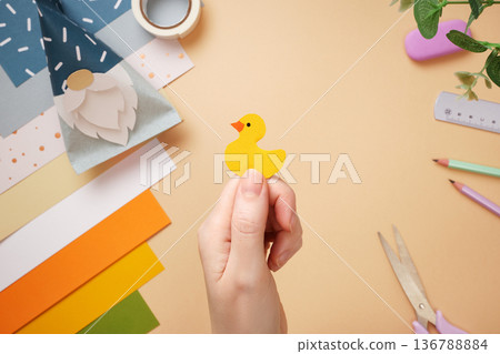 Hands holding yellow paper duck. Ruler, scissors, pencil on beige background. DIY handmade project for children. Selective focus. 136788884