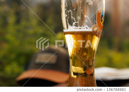 Golden beer glass shining in the sunlight with a casual hat nearby on a warm day outdoors Golden beer glass shining in the sunlight with a casual hat nearby on a warm day outdoors 136788981