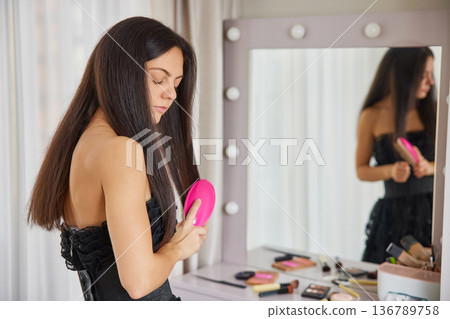 Woman standing in front of illuminated mirror, brushing her long, dark hair 136789758