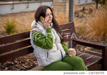 Woman having an outdoor phone conversation while sitting on a bench with coffee 136789795