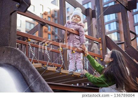 Mother and child playing on a playground bridge, holding hands and smiling 136789804