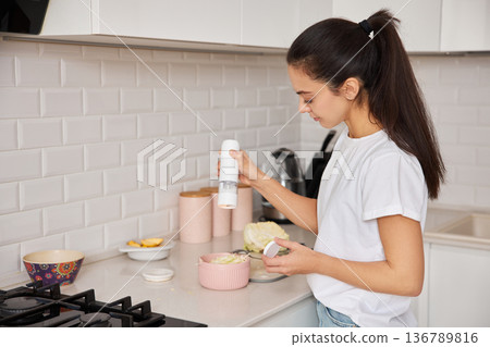 Woman preparing healthy food, adding spice to a meal in a modern kitchen setting 136789816