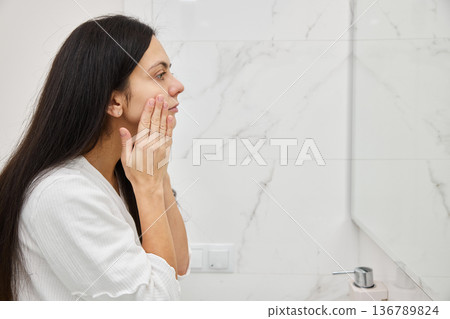 Young woman applying skincare cream to her face in the bathroom mirror performing a beauty routine Young woman applying skincare cream to her face in the bathroom mirror performing a beauty routine 136789824