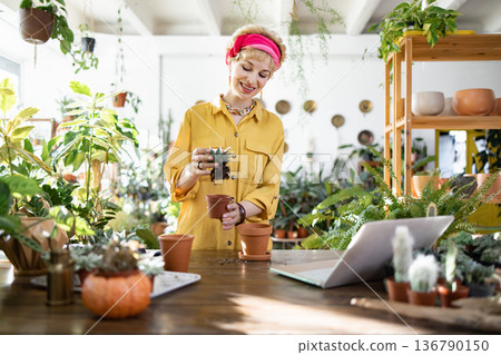 A woman smiles while working on her plants, surrounded by greenery and using a laptop A woman smiles while working on her plants, surrounded by greenery and using a laptop 136790150