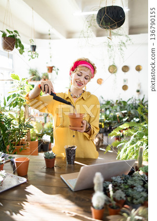 A woman in a yellow shirt and pink headscarf is planting succulents in a terracotta pot, surrounded by lush greenery A woman in a yellow shirt and pink headscarf is planting succulents in a terracotta pot, surrounded by lush greenery 136790155