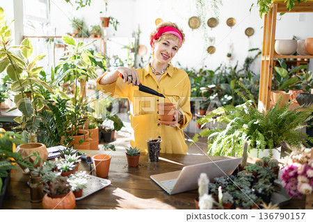 A smiling woman is planting a flower in a pot, surrounded by various plants in a bright, sunny room A smiling woman is planting a flower in a pot, surrounded by various plants in a bright, sunny room 136790157