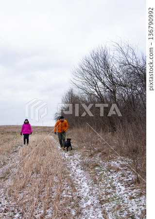 Couple enjoying a chilly winter walk with their dog along a snowy trail bordered by trees in a serene landscape at dusk Couple enjoying a chilly winter walk with their dog along a snowy trail bordered by trees in a serene landscape at dusk 136790992