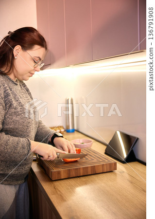 Woman slicing red pepper on wooden cutting board in modern kitchen with tablet on stand. Woman slicing red pepper on wooden cutting board in modern kitchen with tablet on stand. 136791908